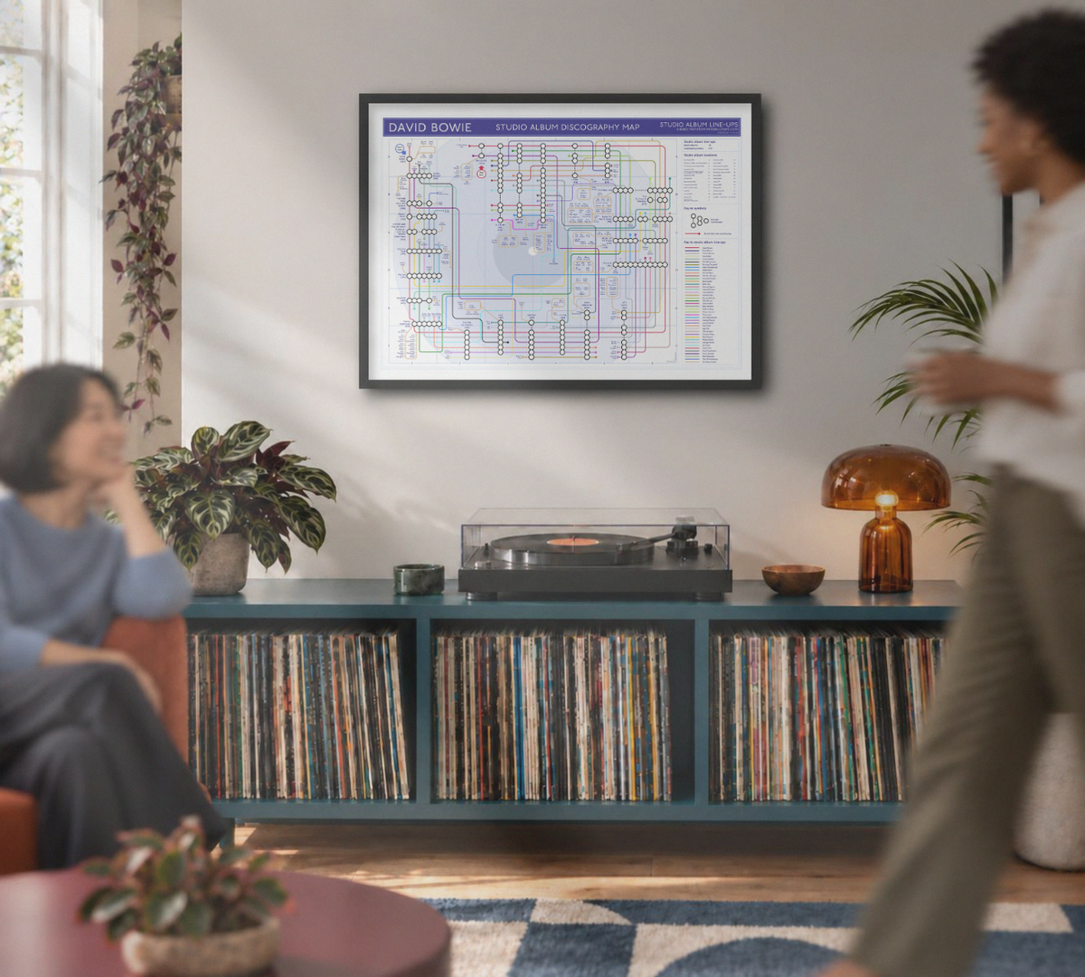 A framed David Bowie print hangs on the wall in a modern living room. The room features a teal record store cabinet filled with vinyl records, a turntable, and plants, with two people visible in the foreground and background.