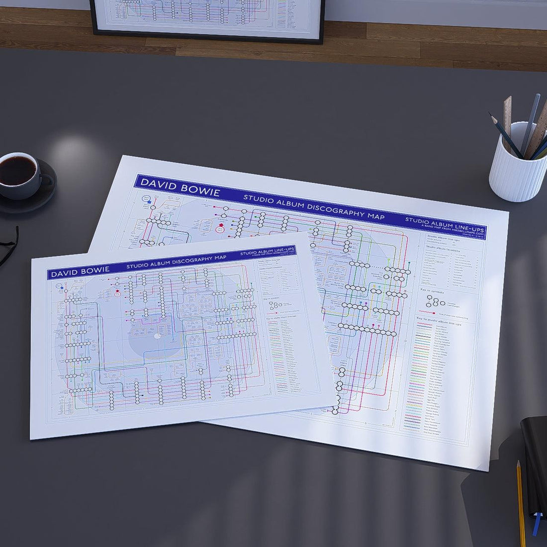 Two David Bowie studio album discography maps on a desk with a cup of coffee and office supplies.